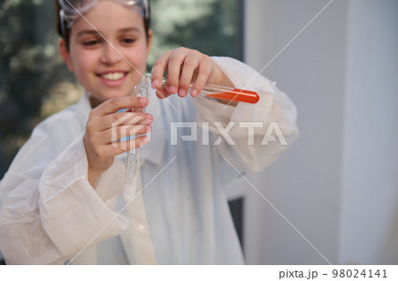 Details on the hands of a blurred Caucasian smiling teenage boy, smart young chemist scientist, in white lab coat, conducting chemical experiment in chemistry laboratory. Kids development. Education Details on the hands of a blurred Caucasian smiling teenage boy, smart young chemist scientist, in white lab coat, conducting chemical experiment in chemistry laboratory. Kids development. Education 98024141