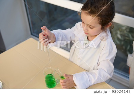 Overhead view of Caucasian little child girl in white lab coat, dripping green chemical liquid into a glass measuring beaker, using graduated pipette in the school chemistry laboratory. Back to school Overhead view of Caucasian little child girl in white lab coat, dripping green chemical liquid into a glass measuring beaker, using graduated pipette in the school chemistry laboratory. Back to school 98024142