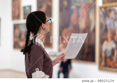Side view of caucasian pretty young woman wearing glasses holds educational brochure. Defocused pictures in background. Concept of exhibition in gallery Side view of caucasian pretty young woman wearing glasses holds educational brochure. Defocused pictures in background. Concept of exhibition in gallery 98025124