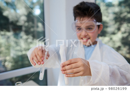 Close-up. Selective focus on hands of blurred teen nerd schoolboy, chemist scientist in safety goggles and white lab coat, holding test tubes with coming out chemical substance. Miracles of Chemistry Close-up. Selective focus on hands of blurred teen nerd schoolboy, chemist scientist in safety goggles and white lab coat, holding test tubes with coming out chemical substance. Miracles of Chemistry 98026199