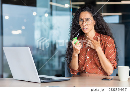 A young sick Latin American woman sits in the office at a table with a laptop, feels bad. He coughs, blows his throat with an aerosol medication. Holds his chest, He looks sadly at the camera. A young sick Latin American woman sits in the office at a table with a laptop, feels bad. He coughs, blows his throat with an aerosol medication. Holds his chest, He looks sadly at the camera. 98026467