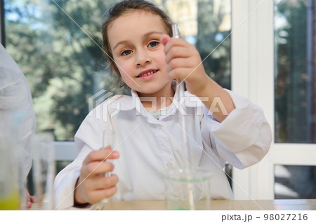 Close-up portrait of a Caucasian blue-eyed lovely little child girl in white lab coat, holding a test tube and a graduated pipette at chemistry class, in laboratory, cutely smiling looking at camera 98027216