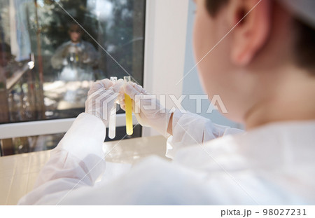 Rear view: selective focus on the test tubes with chemicals in gloved hands of a smart teenage schoolboy in white lab coat, conducting chemical scientific experiment in a chemistry laboratory Rear view: selective focus on the test tubes with chemicals in gloved hands of a smart teenage schoolboy in white lab coat, conducting chemical scientific experiment in a chemistry laboratory 98027231
