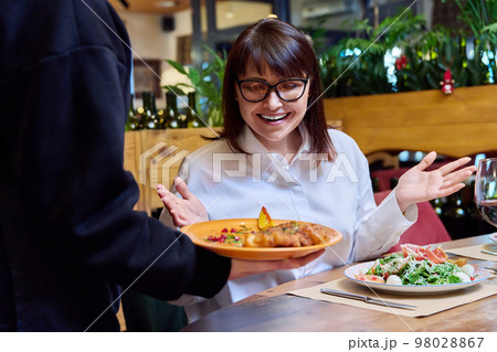 Woman sitting in restaurant, rejoices at plate of cooked food in hands of waiter Woman sitting in restaurant, rejoices at plate of cooked food in hands of waiter 98028867