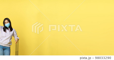 Full length shot of enthusiastic korean girl enjoying vacation, posing with suitcase, wearing face medical mask, travelling abroad during covid pandemic, yellow background Full length shot of enthusiastic korean girl enjoying vacation, posing with suitcase, wearing face medical mask, travelling abroad during covid pandemic, yellow background 98033290