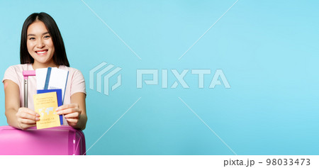 Happy korean woman, tourist with suitcare shows her health passport and tickets, covid-19 international vaccination certificate for travelling, going on trip, blue background 98033473