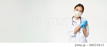 Smiling cute asian female doctor, nurse in medical respirator and rubber sterile gloves, looking pleased at camera, standing in healthcare worker uniform, white background 98033615