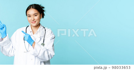 Smiling female doctor, physican in medical uniform and sterile gloves, pointing fingers left at promo, clinic logo, standing over blue background 98033653
