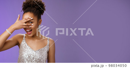 Close-up stylish african american woman afro hairstyle brilliant accessorise glittering silver dress hiding face peeking through fingers amused look surprised anticipating interesting gift 98034050