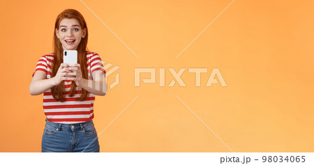 Amused excited redhead female record video thrilled see famous person, smiling amazed hold smartphone taking picture, tourist adore trip shooting everything during summer journey, orange background Amused excited redhead female record video thrilled see famous person, smiling amazed hold smartphone taking picture, tourist adore trip shooting everything during summer journey, orange background 98034065