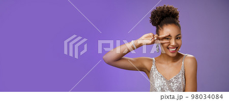 Slim stylish beautiful young elegant african american woman in trendy silver dress brilliant accessorise smiling broadly show peace victory gesture near eye feel lucky amused, blue background 98034084