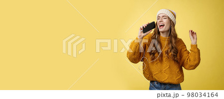 Carefree charming girl likes karaoke having fun listen favorite songs wearing wired earphones hold smartphone microphone singing out loud dancing enjoying spend time alone 98034164