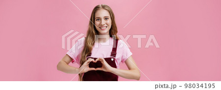 Girl loves family. Kind charming young woman in overalls with small tattoo on arm showing heart gesture over body and smiling lovely at camera expressing tender and cute attitude over pink wall 98034195