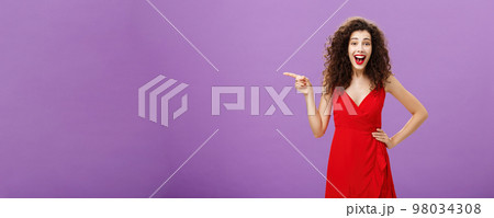 Portrait of entertained and amused joyful european woman with curly hairstyle in red evening dress laughing from amusement and joy pointing left being entertained posing over purple background 98034308