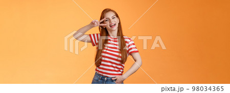 Friendly attractive modern redhead female college star show peace victory sign near eye, smiling happily, express positivity, joy and confidence, motivated win, stand orange background optimistic 98034365