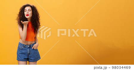 Stylish flirty and confident female with curly hairstyle and red lipstick smiling sensually bending towards camera sending gentle loving kiss at camera with palm near mouth posing near orange wall Stylish flirty and confident female with curly hairstyle and red lipstick smiling sensually bending towards camera sending gentle loving kiss at camera with palm near mouth posing near orange wall 98034469