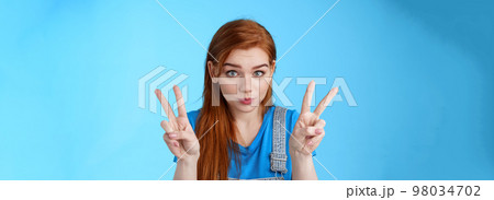 Playful cute silly redhead girlfriend making funny face, show peace victory signs, hold breath, pouting childish, fool around having fun, stand blue background, relaxing entertain siblings 98034702