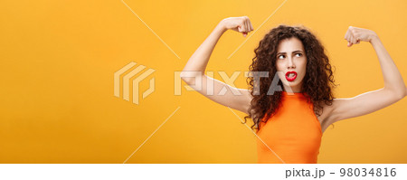 Woman feeling powerful and strong raising hands with clenched fists making intense face being working out in gym showing muscles and biceps looking at upper right corner posing over orange background 98034816