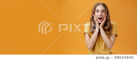 Waist-up shot of surprised and amused girl learning shocking awesome rumor touching cheeks from amazement opening mouth intrigued gazing at camera focused on interesting story over orange background 98034840