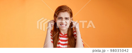 Turn off sound. Annoyed disturbed redhead female cannot study hear bothering loud noise, plug fingers ear holes grimace, clench teeth displeased, sibling awful play violin, orange background 98035042