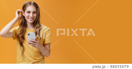 Waist-up shot of cute and friendly-looking nice young girl with wavy hairstyle putting hair strand behind ear holding smartphone smiling broadly with joyful expression at camera over orange background Waist-up shot of cute and friendly-looking nice young girl with wavy hairstyle putting hair strand behind ear holding smartphone smiling broadly with joyful expression at camera over orange background 98035079