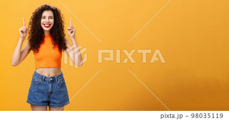 Indoor shot of friendly and cute happy relaxed female with red lipstick and curly hairstyle wearing cool cropped top and denim shorts smiling broadly pointing up posing over orange background Indoor shot of friendly and cute happy relaxed female with red lipstick and curly hairstyle wearing cool cropped top and denim shorts smiling broadly pointing up posing over orange background 98035119