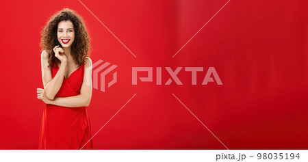 Passionate charming european woman over red background in elegant dress with curly hairstyle smiling cute, feminine playing with hair strand standing timid and silly, talking to person she admires Passionate charming european woman over red background in elegant dress with curly hairstyle smiling cute, feminine playing with hair strand standing timid and silly, talking to person she admires 98035194
