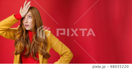 Redhead businesswoman making facepalm gesture with hand on forehead rolling eyes up from annoyance and irritation as being shocked with how dumb client is sighing bothered over red wall, tired 98035220