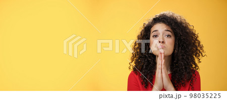 Please I beg you. Portrait of sad and cute curly-haired girl with angel eyes pouting holding hands in pray and looking hopeful at camera waiting for mercy asking apology, supplicating over yellow wall 98035225