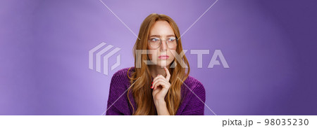 Girlfriend solving puzzle like detective making assumptions focusing on problem squinting looking left determined and thoughtful wearing glasses touching jaw while thinking over purple background 98035230