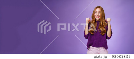 Yey we made it. Happy and cheerful young pretty redhead 20s girl in purple sweater over violet background raising clenched fists in joy and triumph, celebrating success and win, cheering 98035335