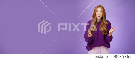 What wrong, I worried. Portrait of nervous and empathical young redhead woman feeling anxious raising hands questioned and looking at camera wondered expressing concern for friend over purple wall 98035366