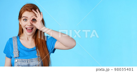 Dreamy curious attractive cheerful redhead woman surprised, look fascinated camera look through fingers okay sign, stand amused, gaze admiration blue background, check out promo 98035401