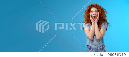 Amazed gasping shocked wondered redhead attractive curly girl popping eyes stare camera drop jaw concerned astonished touch cheeks empathy amazement standing blue background Amazed gasping shocked wondered redhead attractive curly girl popping eyes stare camera drop jaw concerned astonished touch cheeks empathy amazement standing blue background 98036330
