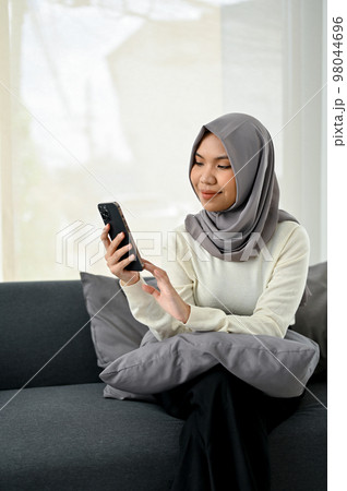 Charming young Asian Muslim woman wearing hijab using her smartphone while relaxing on sofa Charming young Asian Muslim woman wearing hijab using her smartphone while relaxing on sofa 98044696
