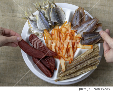 Dried Seafood Mix with Squid, Mussels, Tuna, Yellow Minke, Dried Caviar. Top View. Cured Dehydrated Fish Fillet as Beer Snack Nice Serving. In White Round Plate. Company of people take an appetizer. 98045285