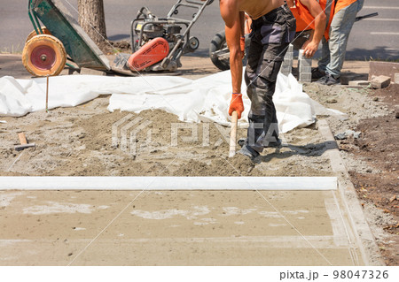 A team of workers with a shovel levels the sandy foundation under the aluminum level for the subsequent laying of paving slabs. 98047326