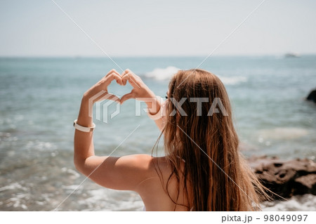 Woman makes heart with hands on beach. Young woman with long hair in white swimsuit and boho style braclets practicing by sea ocean. Concept of longing daydreaming love peace contemplation vacations 98049097