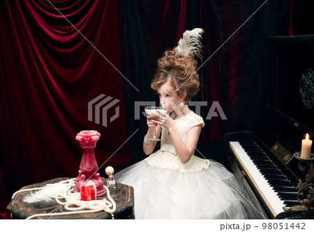 Portrait of cute little girl, child in image of medieval royal person sitting at the piano and drinking milkshake 98051442