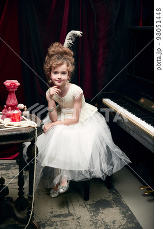 Portrait of cute little girl, child in image of medieval royal person in fabulous dress sitting at the piano and posing 98051448