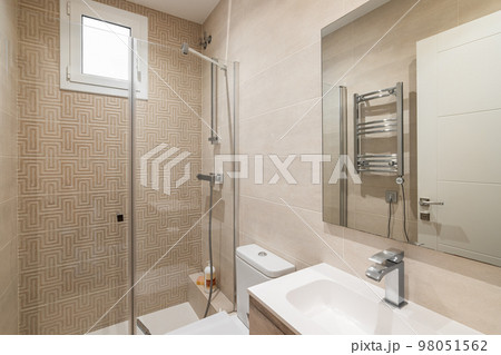 Bright bathroom in beige colors with glossy ceramic tile walls and lighting. The bathroom has a shower with a glass railing, a large mirror with a sink, a radiator for drying towels. Bright bathroom in beige colors with glossy ceramic tile walls and lighting. The bathroom has a shower with a glass railing, a large mirror with a sink, a radiator for drying towels. 98051562