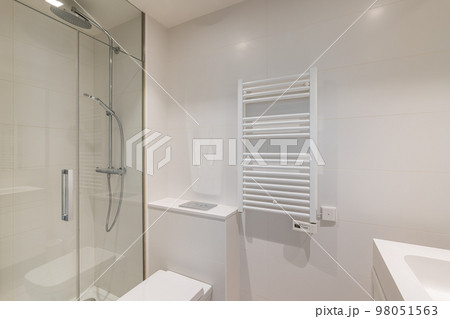Bathroom in light colors with glossy ceramic tile walls and bright lighting. The bathroom has a shower with a glass railing and radiator for drying towels. Bathroom in light colors with glossy ceramic tile walls and bright lighting. The bathroom has a shower with a glass railing and radiator for drying towels. 98051563