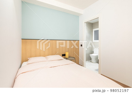 Double bed against a shallow niche with a blue wall and wooden decor. In the bedroom, there is an open door overlooking a bright toilet room with a toilet bowl and a small plastic window. 98052197