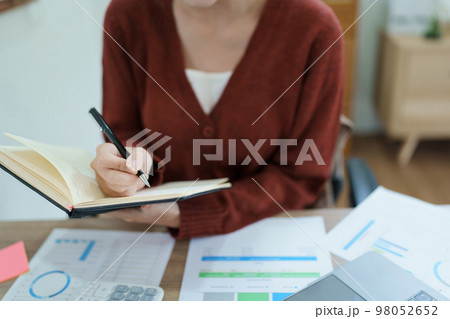 Portrait of a thoughtful Asian businesswoman looking at financial statements and making marketing plans using a computer and notebook on her desk. Portrait of a thoughtful Asian businesswoman looking at financial statements and making marketing plans using a computer and notebook on her desk. 98052652