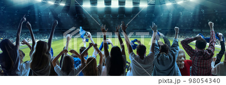 Back view of football, soccer fans emotionally cheering their team at crowded stadium at evening time. Diverse group. Concept of sport, hobby, leisure time, football 98054340