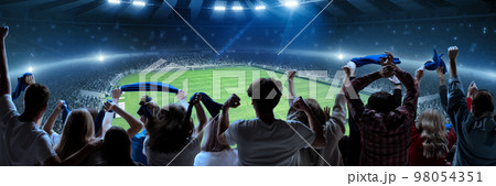 Back view of football, soccer fans cheering their team, holding flag at crowded stadium at evening time. Burst of emotions. Concept of sport, hobby, leisure time, football Back view of football, soccer fans cheering their team, holding flag at crowded stadium at evening time. Burst of emotions. Concept of sport, hobby, leisure time, football 98054351