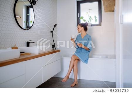 Young woman in a bathroom with phone and cup of coffee, morning routine and relaxation concept. Young woman in a bathroom with phone and cup of coffee, morning routine and relaxation concept. 98055759