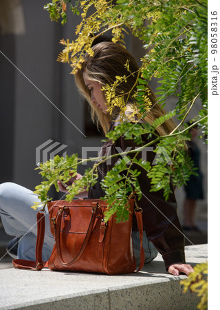 photo of sitting beautiful woman in a blue jeans and brown jacket with orange leather bag 98058316
