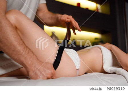 Masseur doing massage of woman's thighs. Masseur at spa salon using gua sha metal scrapper to relieve muscle tension of female body Masseur doing massage of woman's thighs. Masseur at spa salon using gua sha metal scrapper to relieve muscle tension of female body 98059310