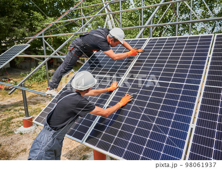 Workers installing photovoltaic solar panel system outdoors. Men engineers placing solar module on metal rails, wearing construction helmets and work gloves. Renewable and ecological energy. Workers installing photovoltaic solar panel system outdoors. Men engineers placing solar module on metal rails, wearing construction helmets and work gloves. Renewable and ecological energy. 98061937